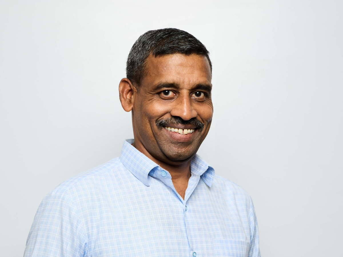 Mr D S Sakthivel in a light blue checkered shirt smiling with hands in pockets against a light grey wall